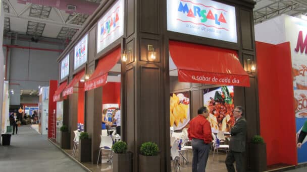 Estande da Marsala montado pela LXBR em feira de alimentos, com toldos bistrô vermelhos, painéis de pratos e área de degustação em madeira.